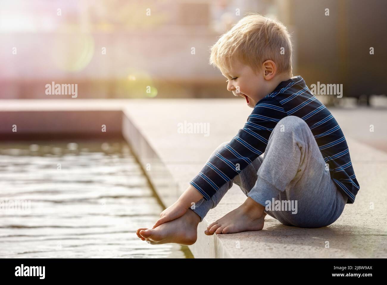 A hilarious boy on a hot summer day in the city is happy to try the cool water of the fountain on his barefoot. The child is surprised by the low wate Stock Photo