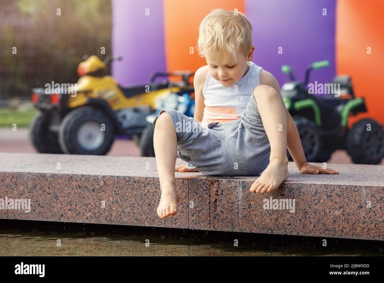 A little barefoot boy in the city plays near the fountain with water at summer. Stock Photo
