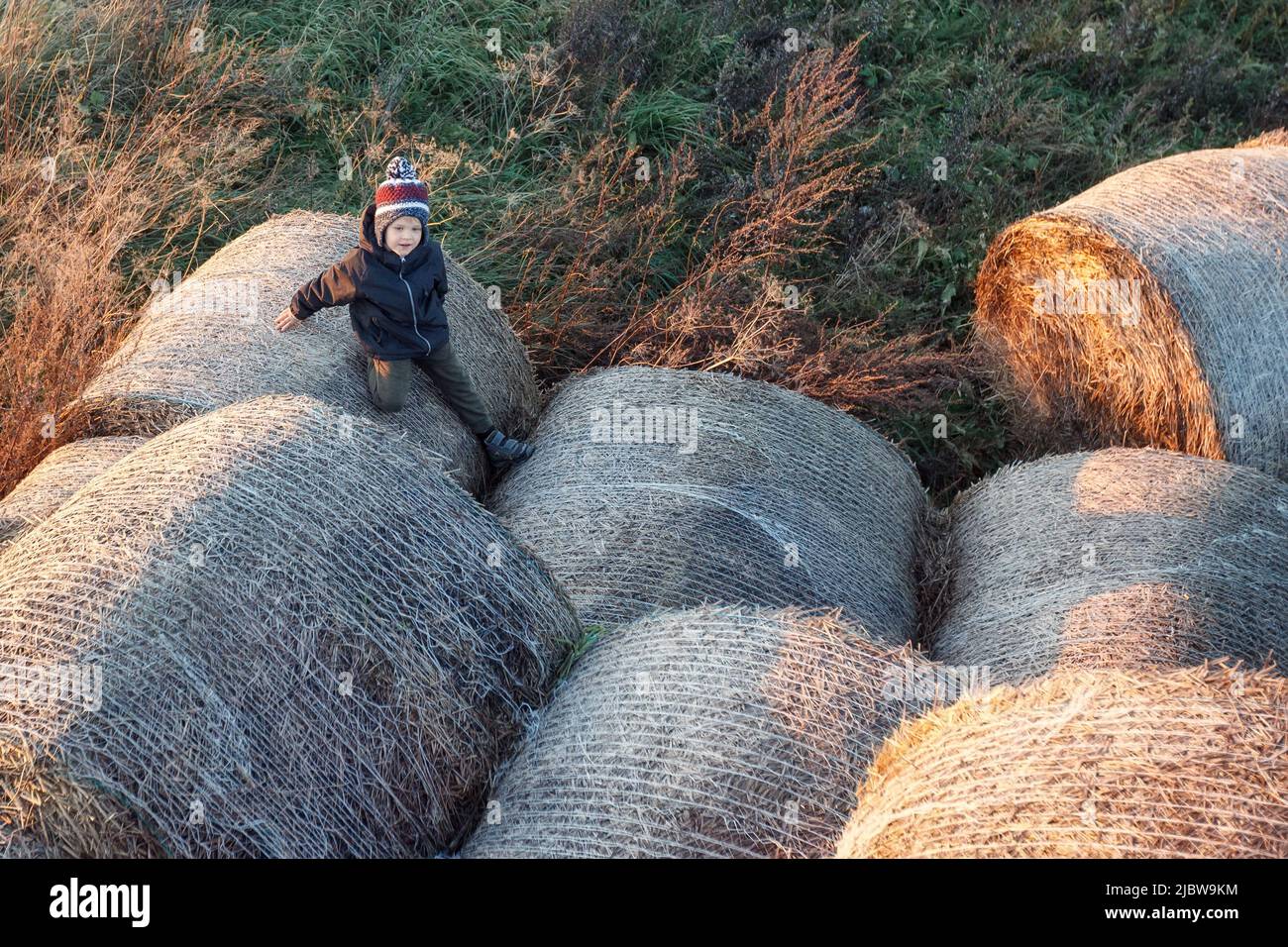 A little cute boy with a warm autumn hat among large rolls of straw in the evening sunlight, photo taken from above. Stock Photo