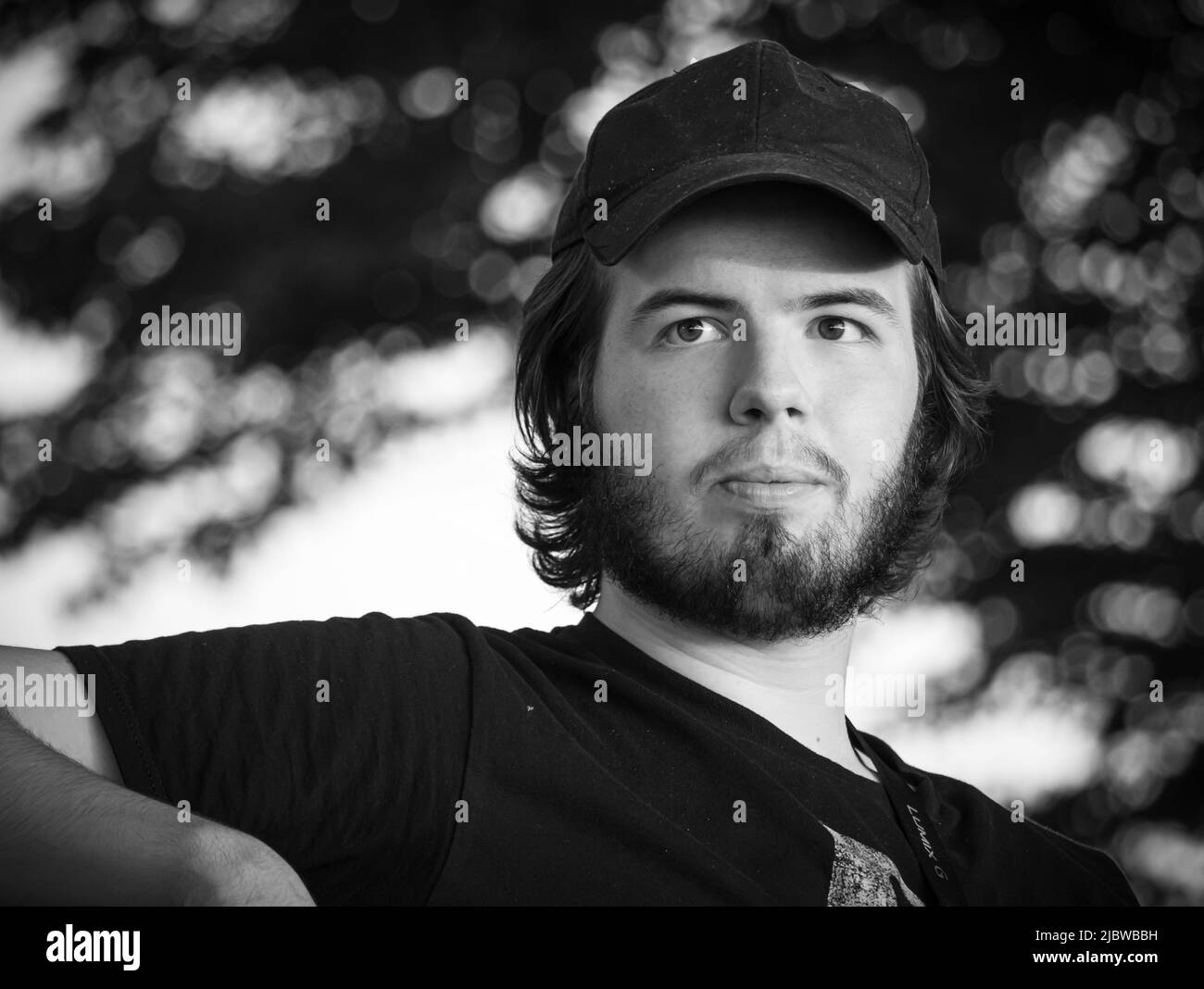 Young adult man with longer dark hair, dark eyes, dark beard,and moustache with a baseball cap sitting at the park contemplating in summer Stock Photo