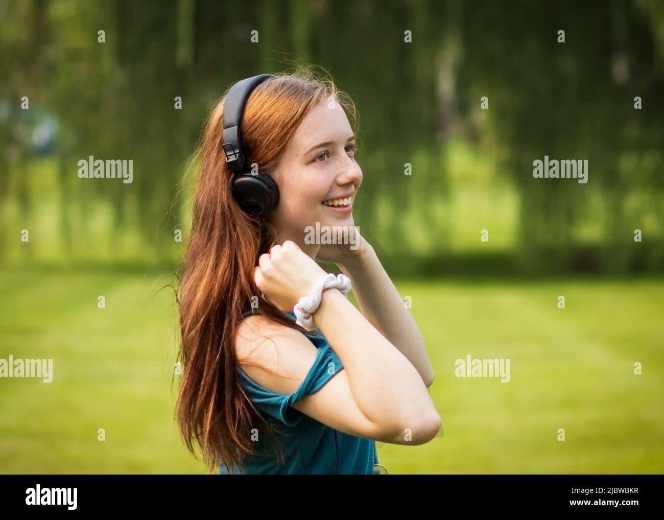 Teenage girl with long red hair smiling and wearing headphones in a green field with a willow tree and green lawn in the background during the summer Stock Photo
