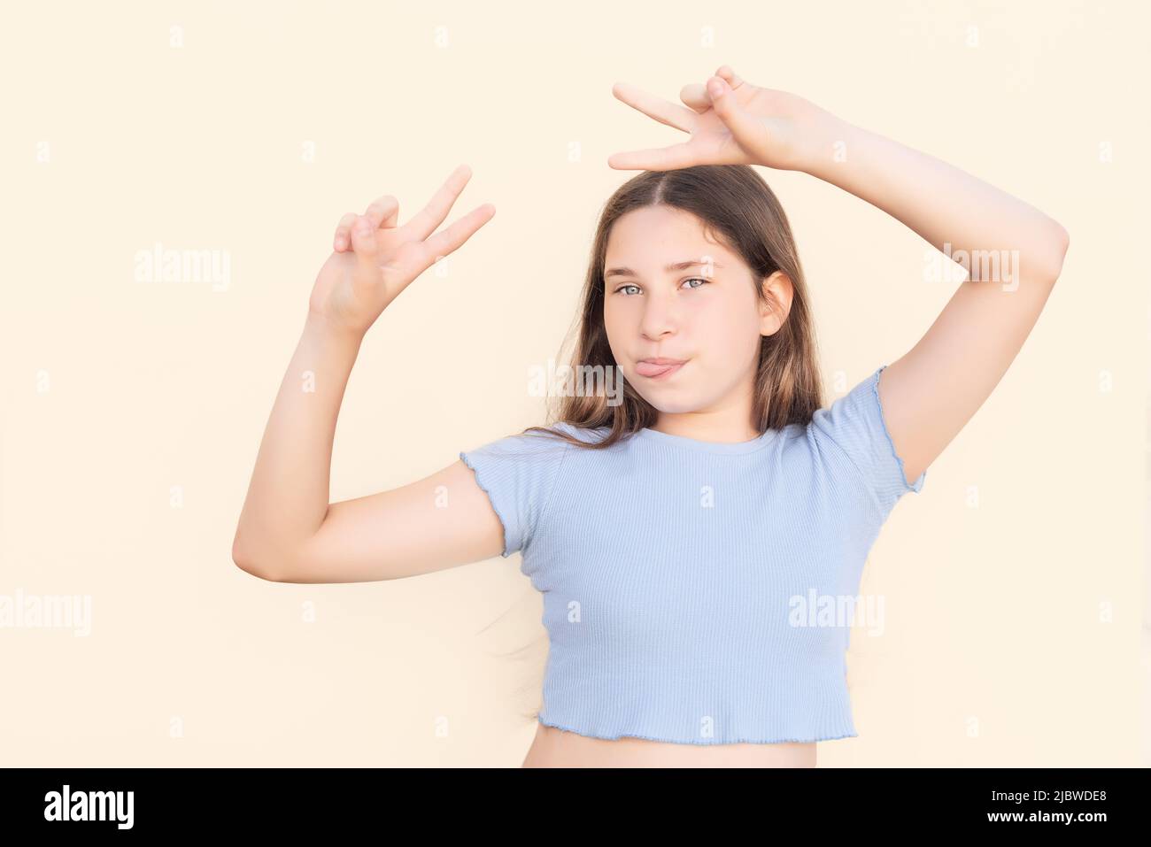 Young Caucasian girl making funny gesture with tongue out and fingers in victory sign on beige isolated background Stock Photo