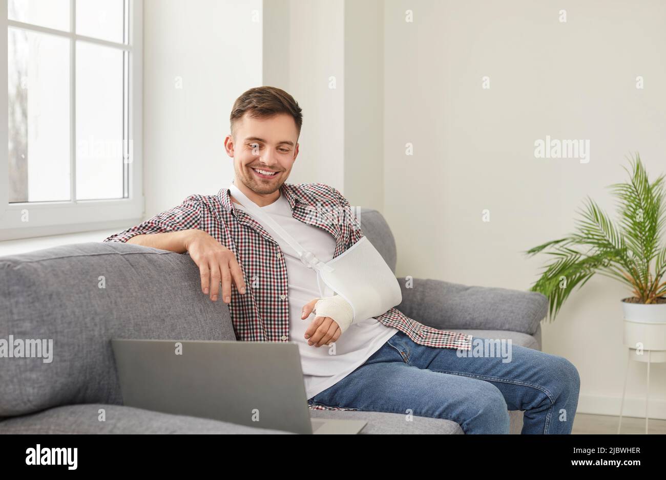 Happy young man with broken arm sitting on sofa at home and watching movie on laptop Stock Photo