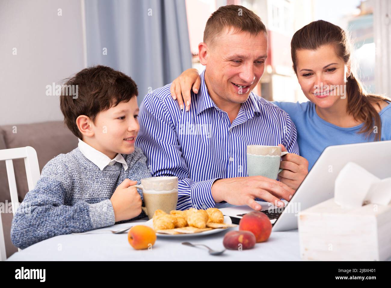 Happy ordinary couple with teenager son using laptop during breakfast at home Stock Photo