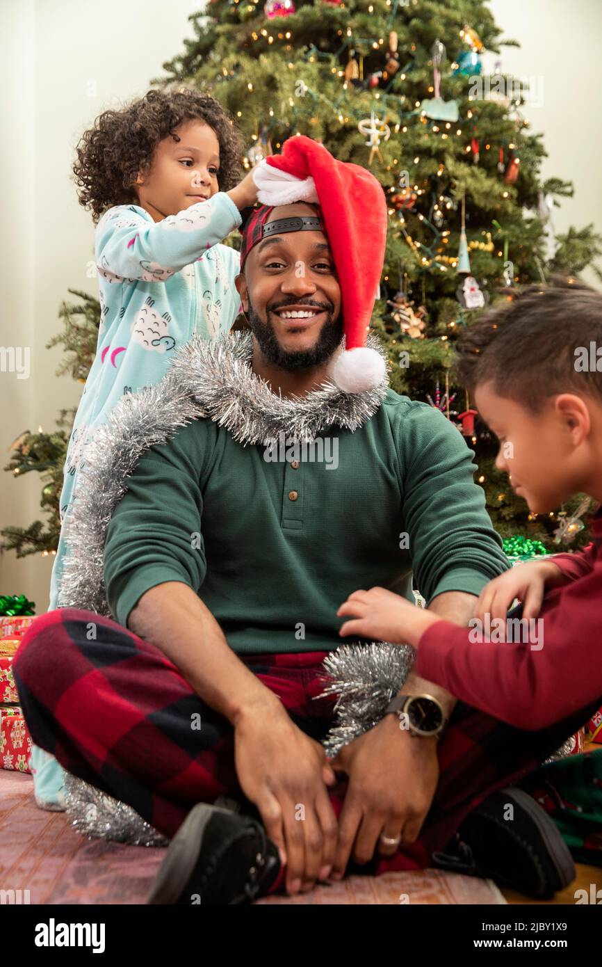 Man sitting in front of Christmas tree being decorated by his children Stock Photo