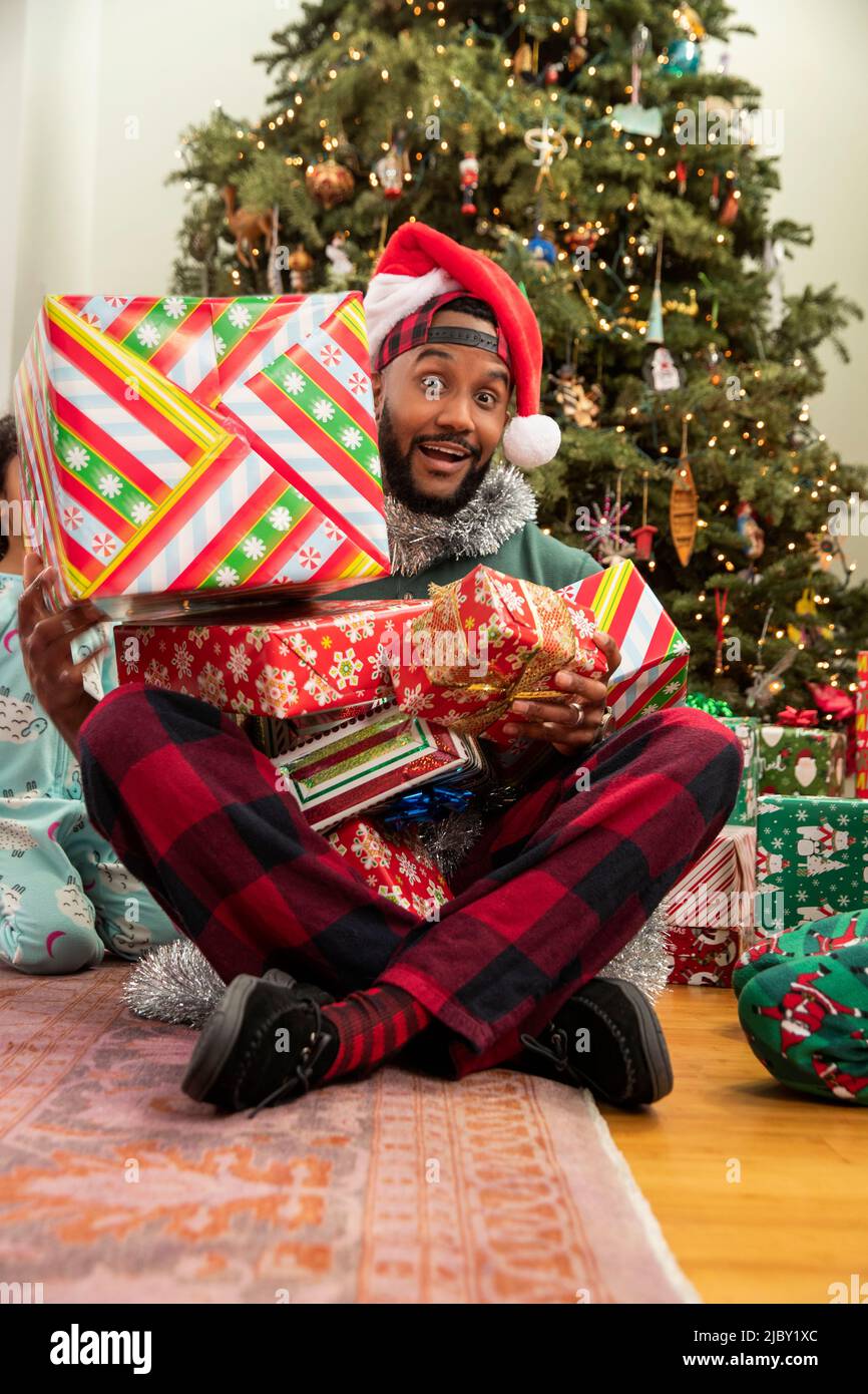 Portrait of father sitting in front of Christmas tree with his kids around him with pile of wrapped presents in his lap. Stock Photo