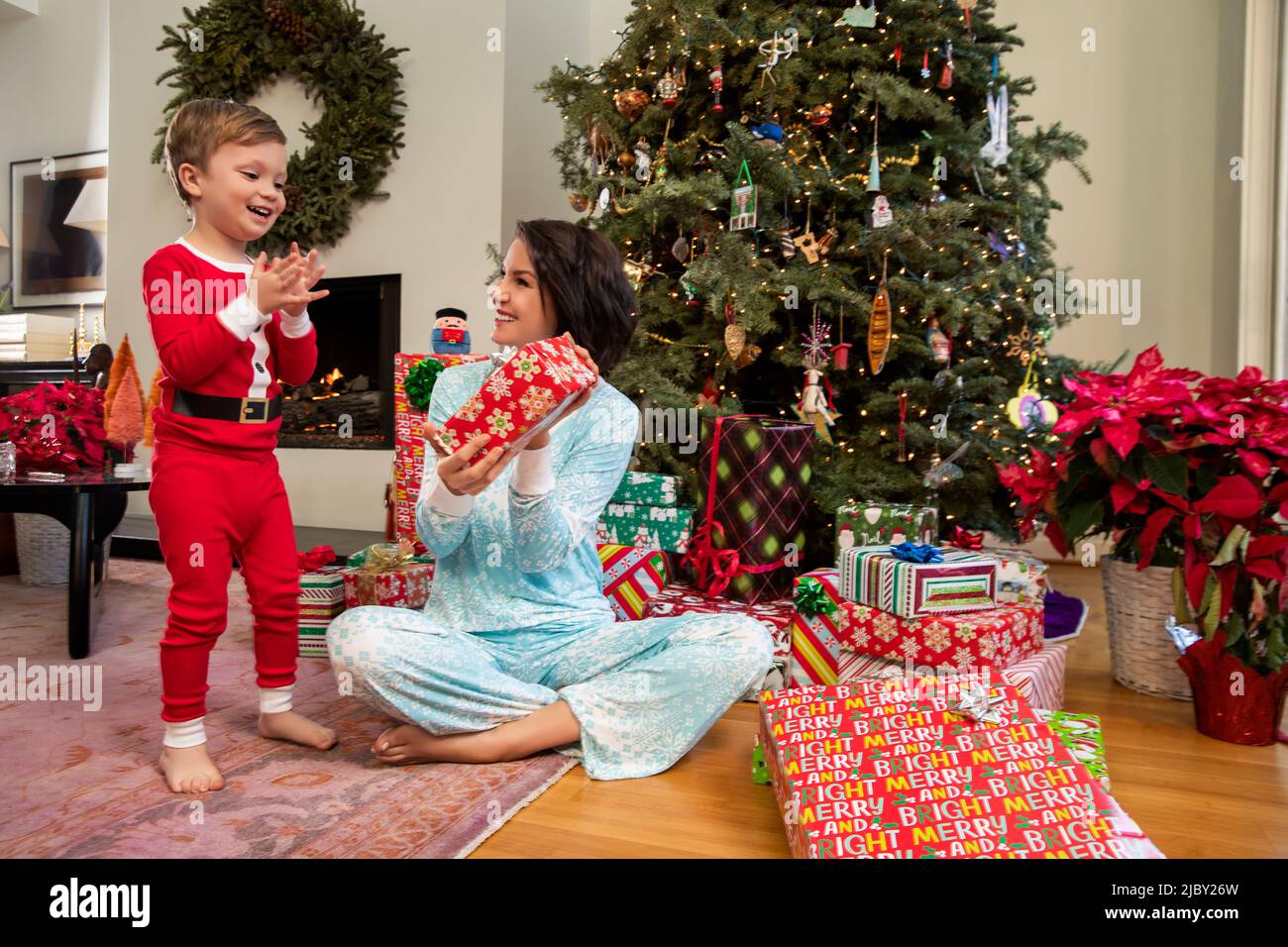 Boy in Santa pajamas clapping hands in excitement as mother hands him Christmas gift Stock Photo