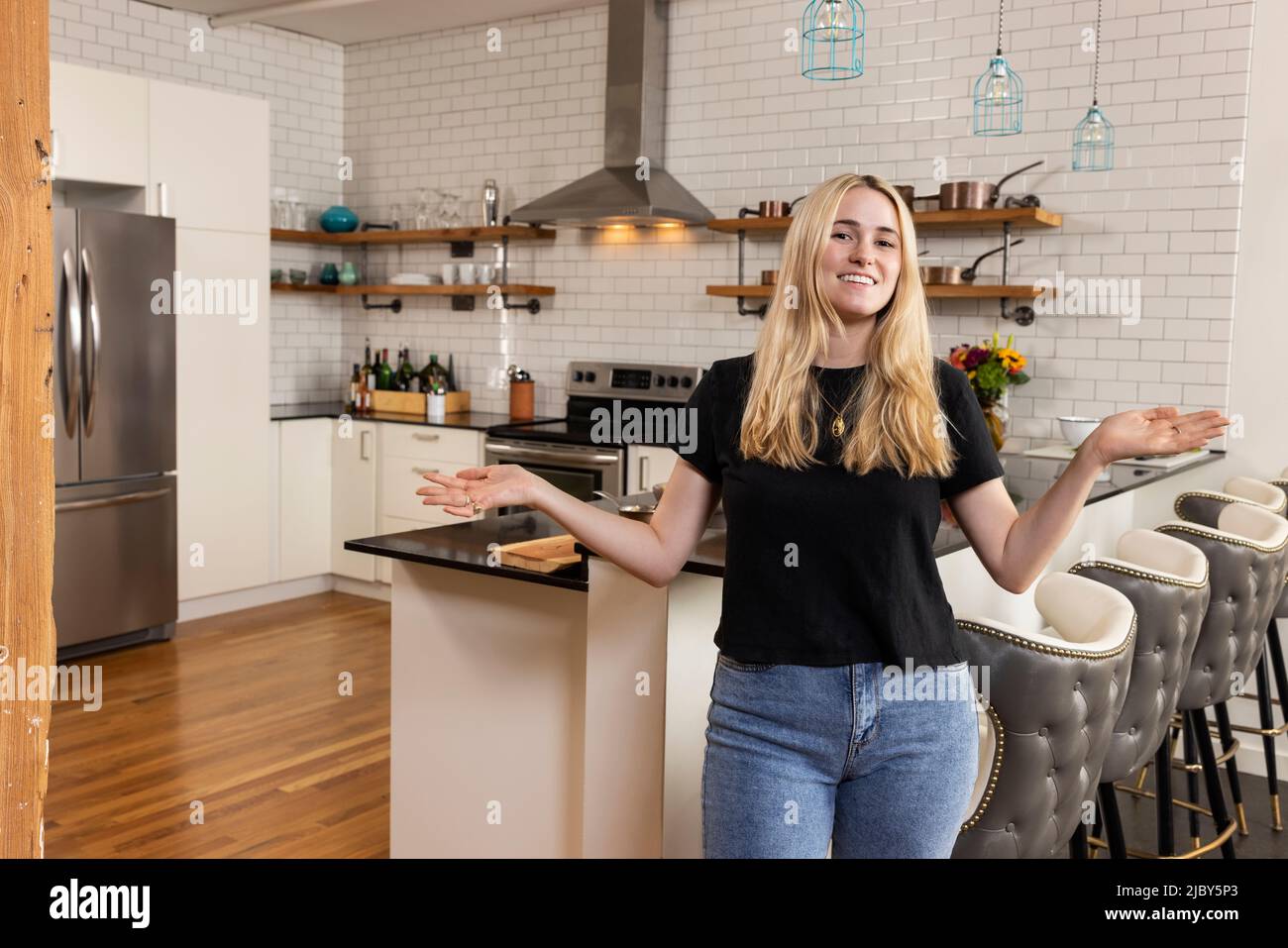 Portrait of a young woman standing in her home kitchen looking at camera with her arms out in a welcoming manner. Stock Photo