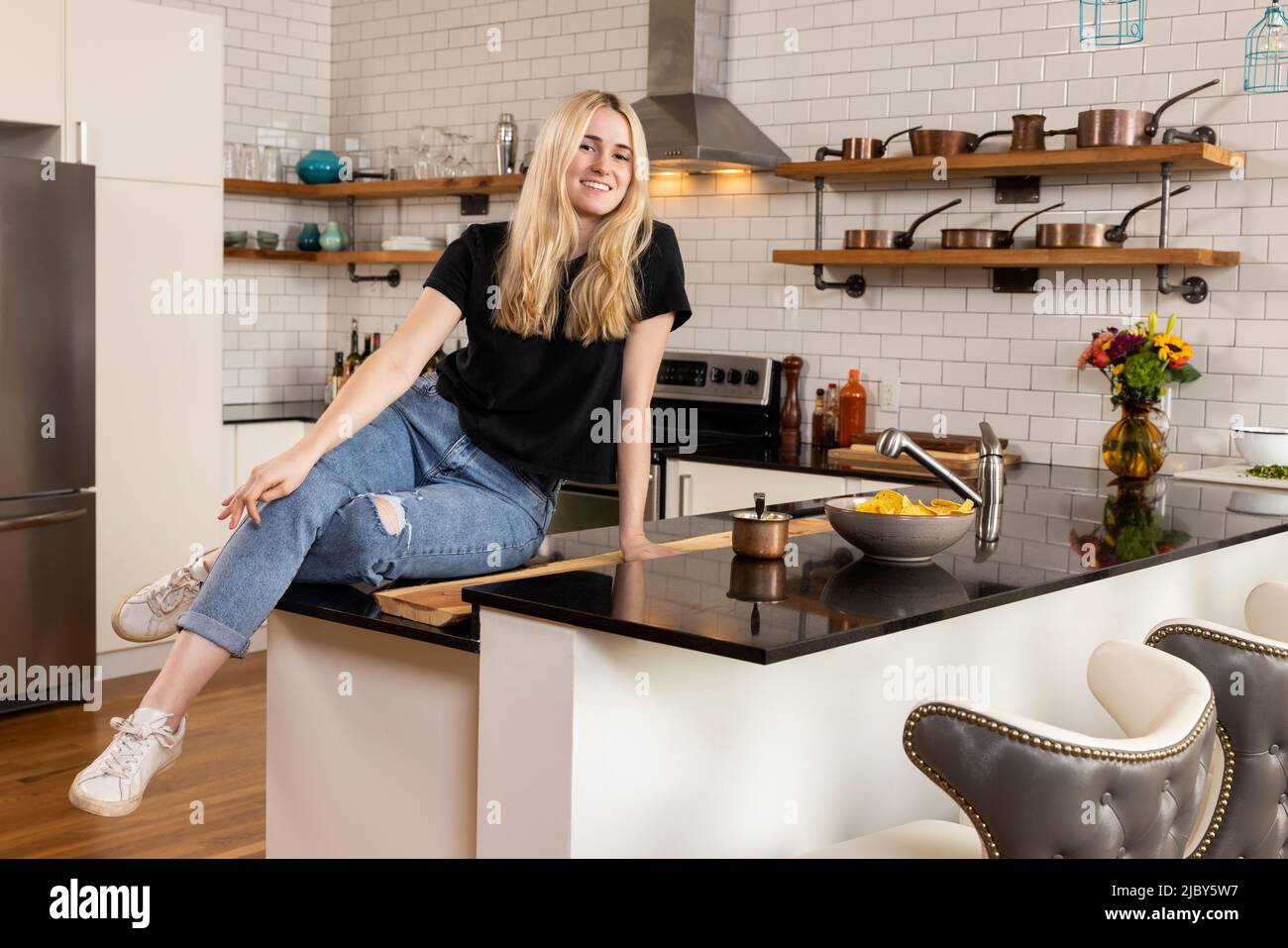 Portrait of smiling young Caucasian woman looking at camera smiling, while sitting on the countertop of her apartment kitchen. Stock Photo
