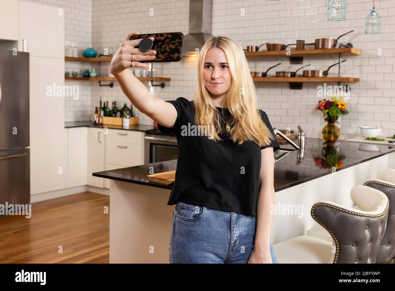 Young adult woman taking selfie while standing in her new apartment with kitchen in background. Stock Photo