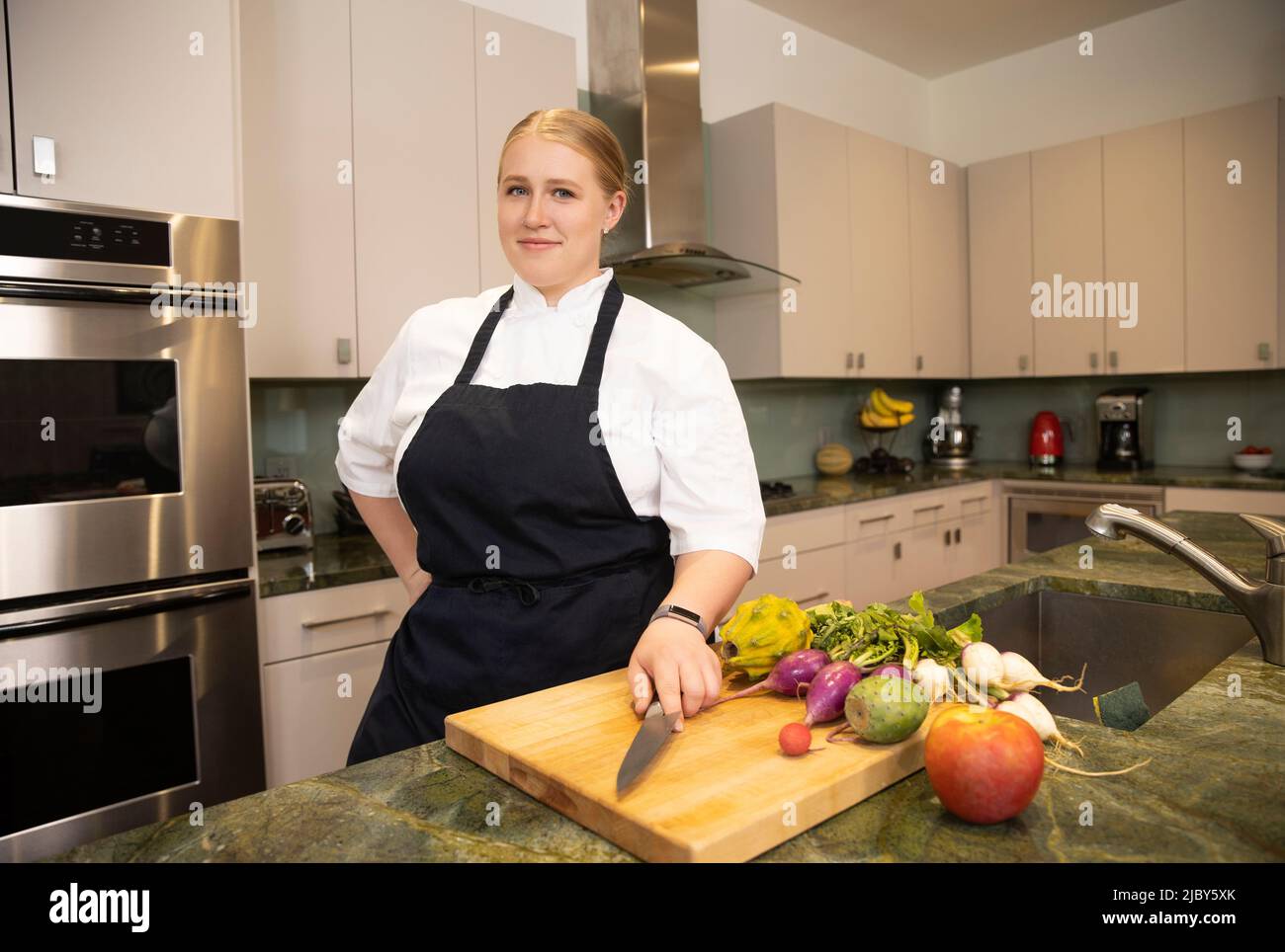 Chef Megan Gill in a kitchen looking at camera with confidence, while having one hand on her culinary knife. Stock Photo