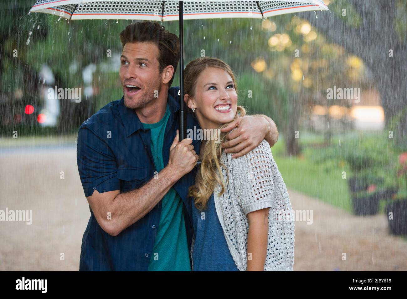 Young couple walking through park with umbrella in the rain, both looking out at the falling rain Stock Photo