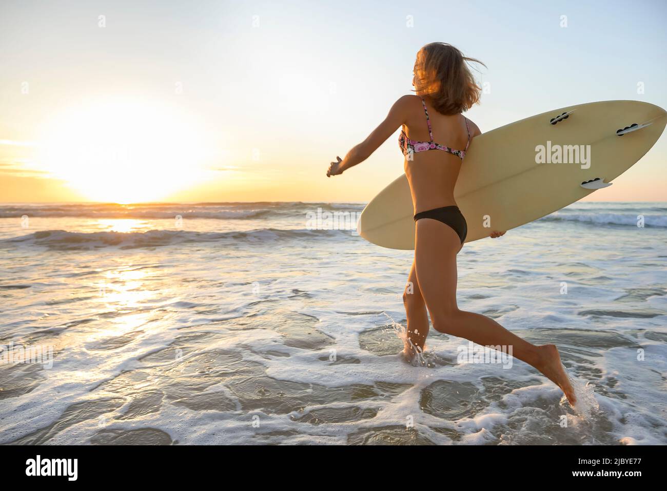 Young woman entering onshore waves holding surfboard in early morning Stock Photo