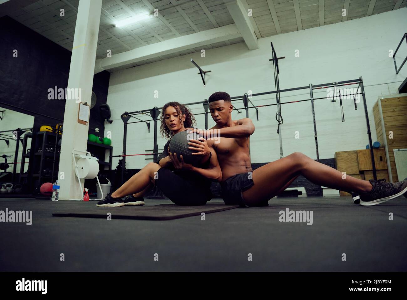 Mixed race friends doing cross fit in the gym. African American male and female sitting down on the floor and passing a medicine ball during exercise Stock Photo
