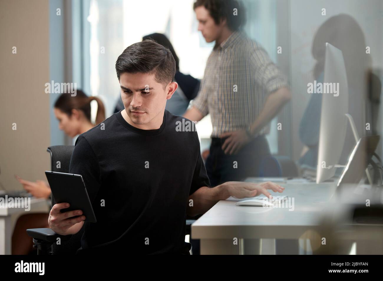 Young man multi-tasking by sitting at computer in office and checking tablet, co-workers having discussion around dry erase board in background Stock Photo