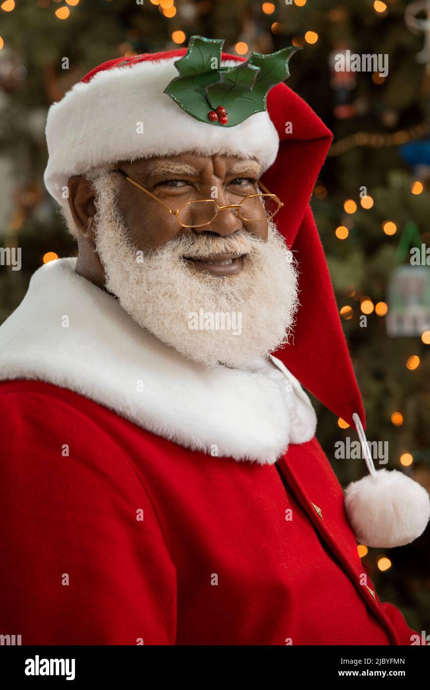 An African American man dressed as Santa Claus sitting in front of a Christmas tree looking camera smiling. Stock Photo