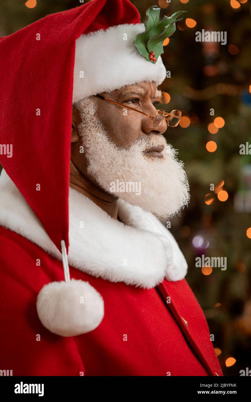 An African American man dressed as Santa Claus sitting in front of a Christmas tree turned profile, looking off camera. Stock Photo