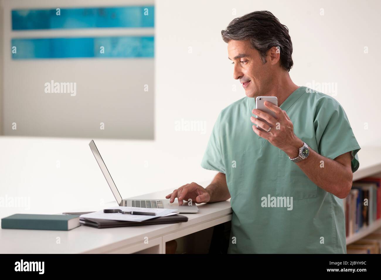 Hispanic Male doctor in his home practicing Telemedicine, talking on cell phone and using laptop computer Stock Photo