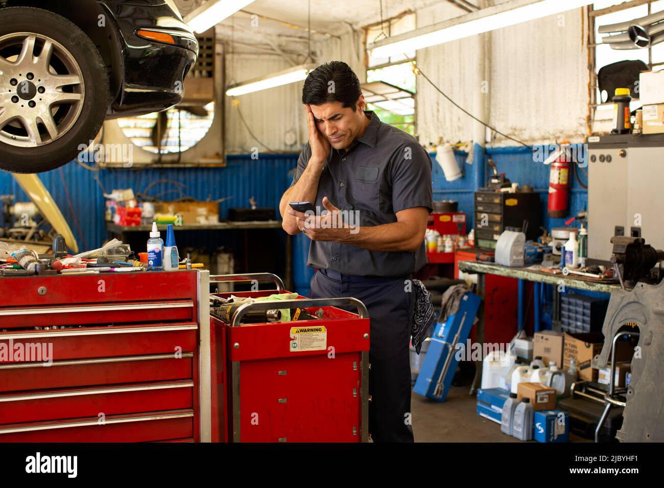 Hispanic car mechanic working in auto shop, feeling ill, using mobile
