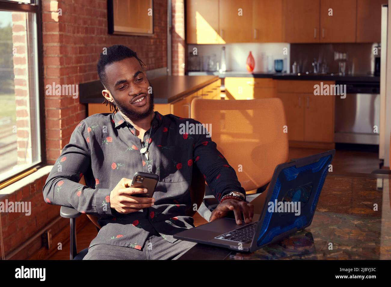 portrait of young ethnic man sitting at conference table using smartphone and laptop computer. Stock Photo