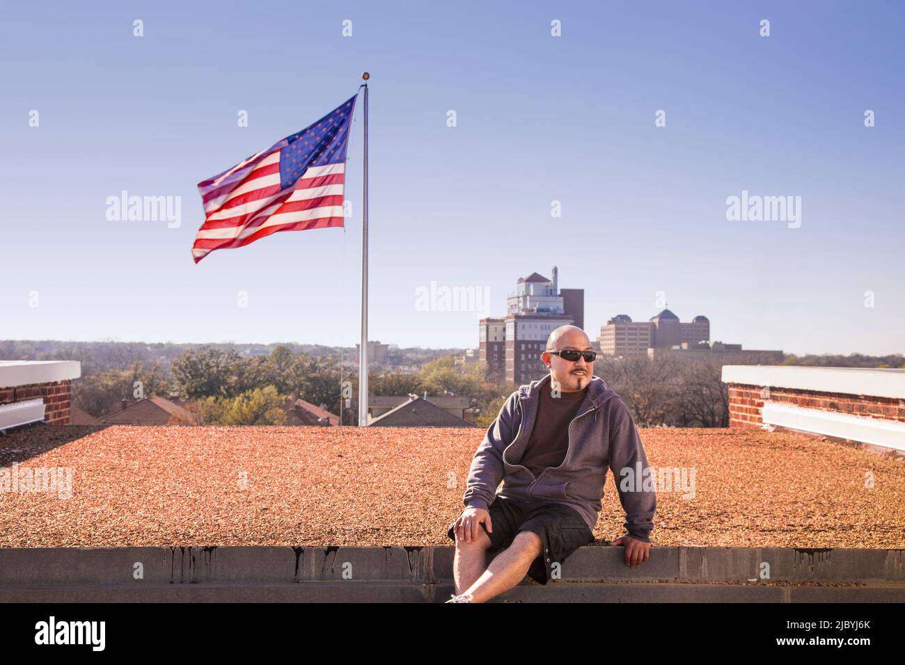 Smiling man sitting on rooftop near American flag Stock Photo
