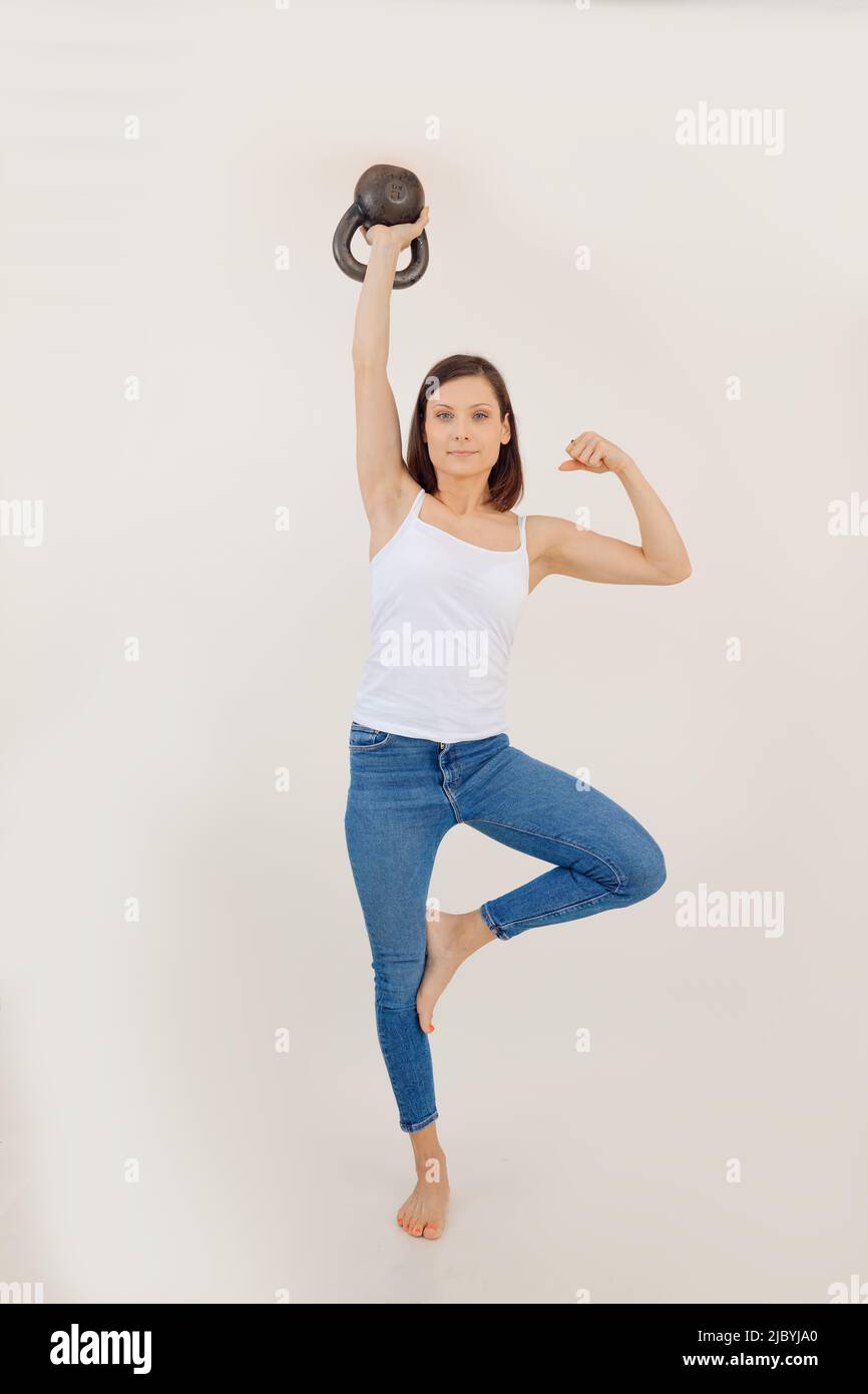 Portrait of young muscular woman wearing white top, blue jeans, standing on leg, lifting kettle bell, showing muscles. Stock Photo