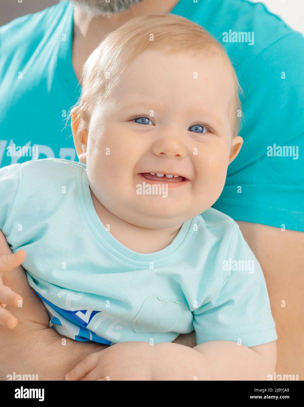 Portrait of family wearing blue T-shirts. Little smiling blue-eyed baby toddler child with fair hair sitting on man. Stock Photo