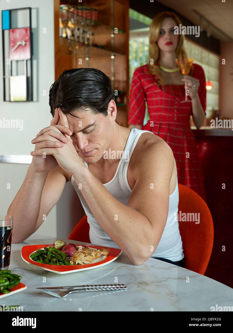 Portrait of a man sitting at a dining table with his wife over his shoulder, looking frustrated. Stock Photo