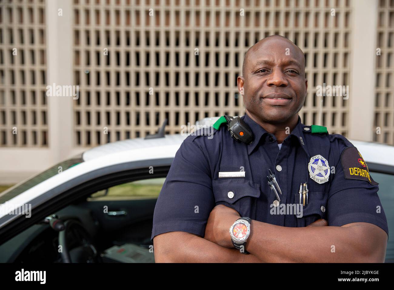 Portrait of Police officer Standing in street in front of squad car looking towards camera with arms behind his back Stock Photo