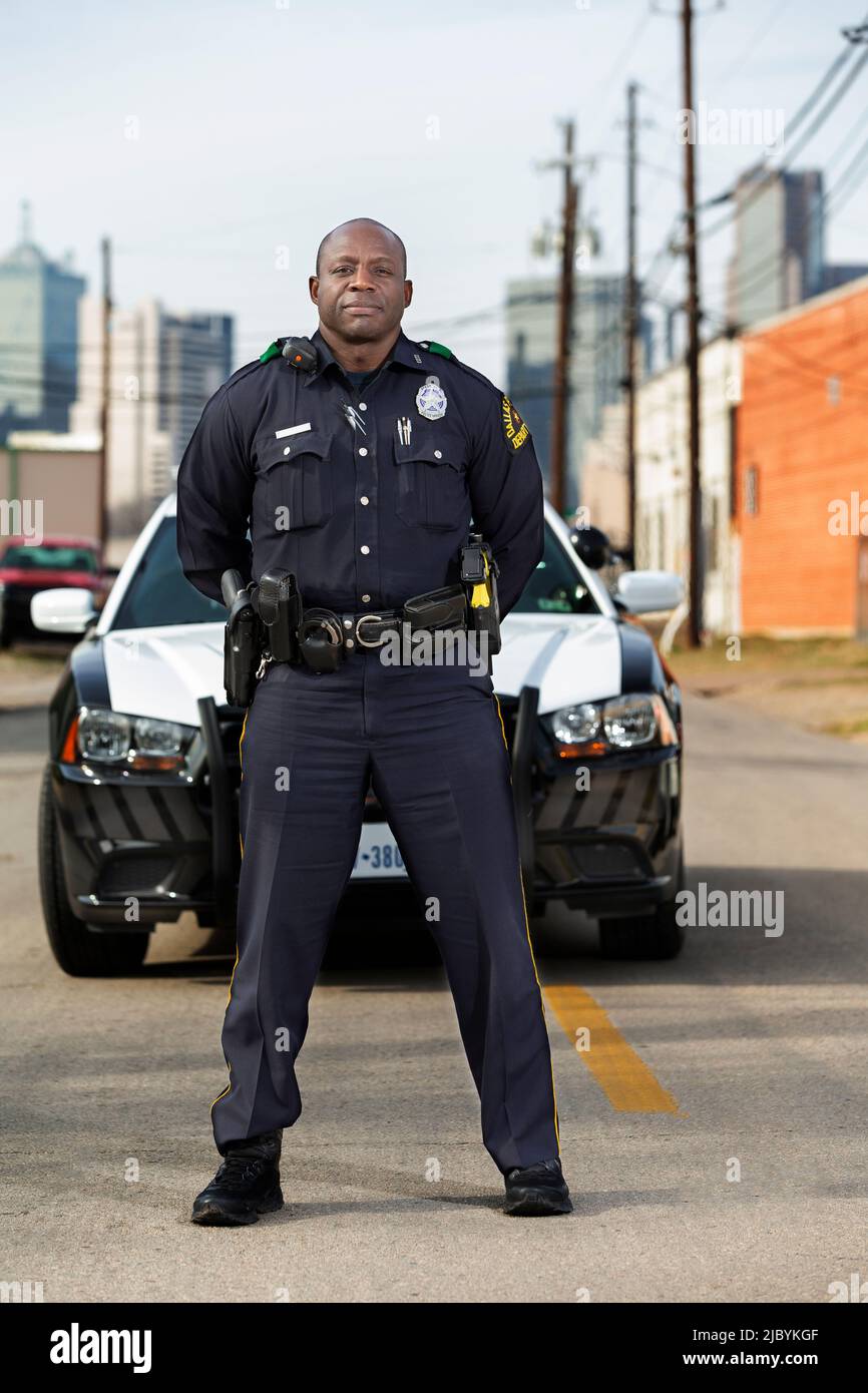 Portrait of Police officer Standing in street in front of squad car looking towards camera with arms behind his back Stock Photo