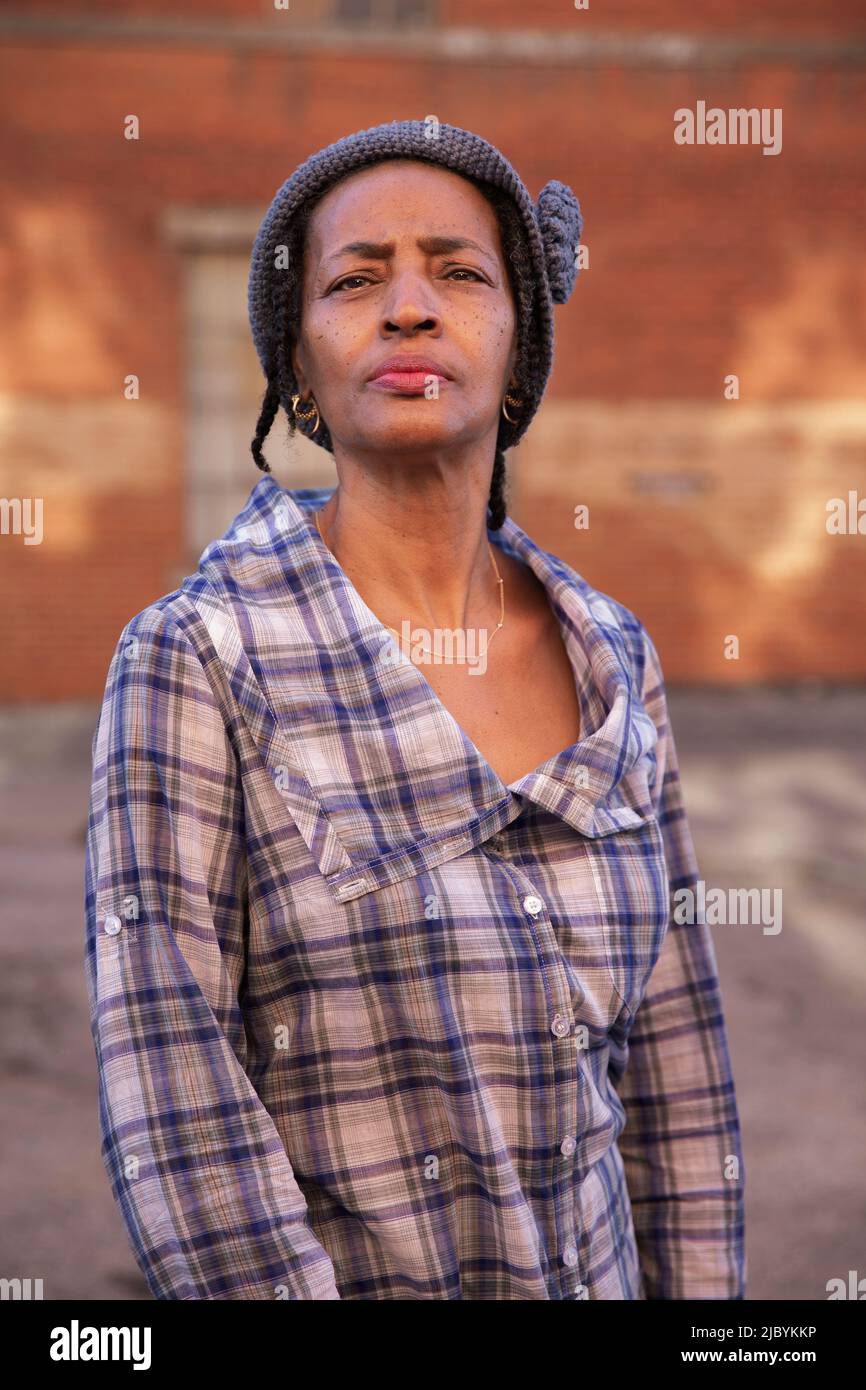 Portrait of older woman wearing knit hat standing in alley holding head up high, brick wall in background Stock Photo