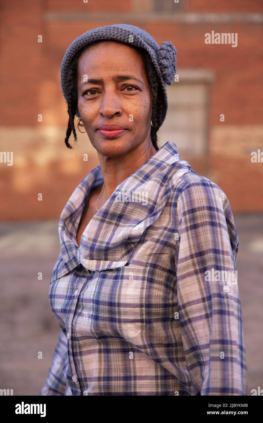Portrait of older woman wearing knit hat standing in alley, brick wall in background Stock Photo