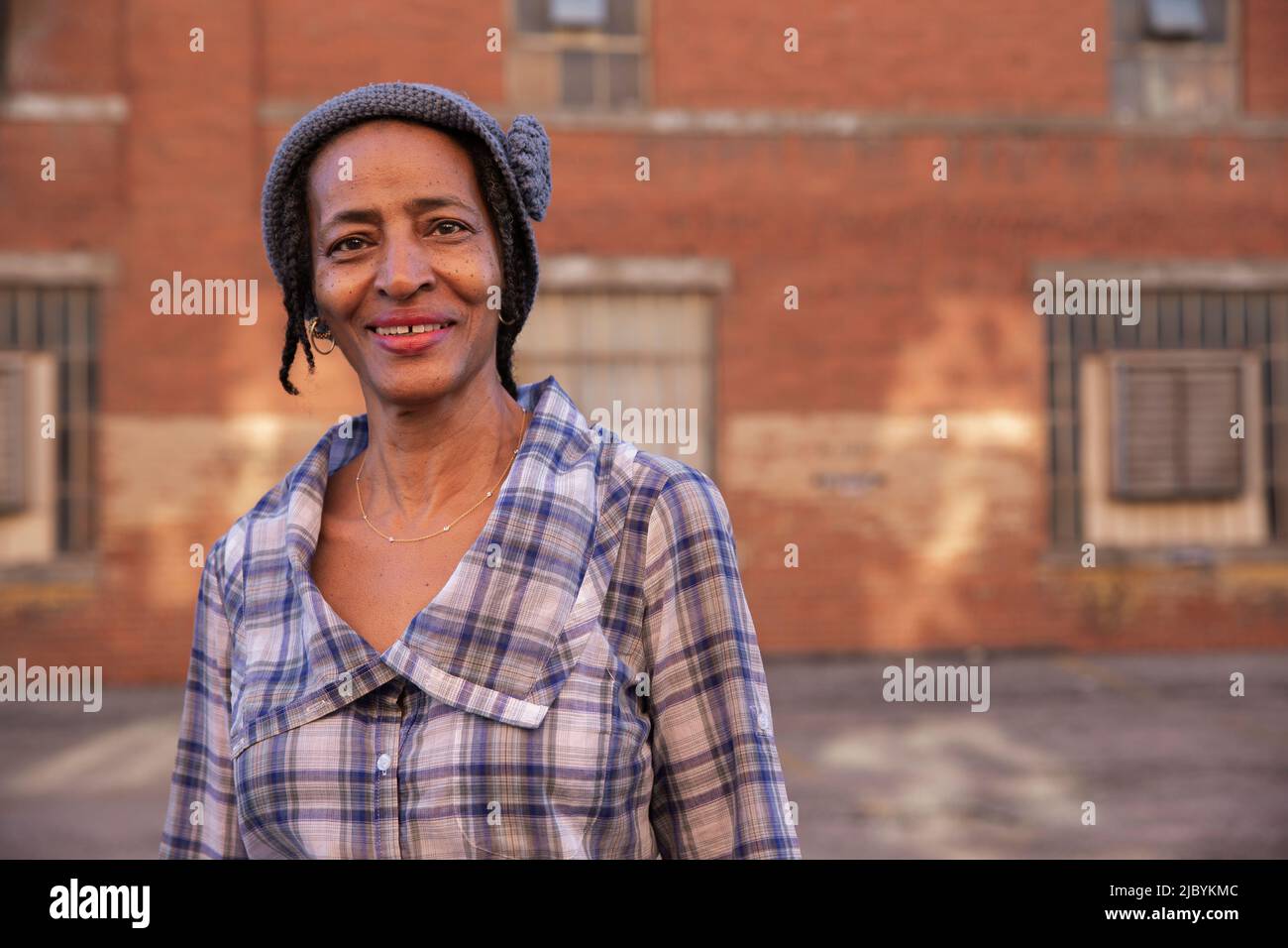 Portrait of older woman wearing knit hat standing in alley smiling looking at camera, brick wall in background Stock Photo