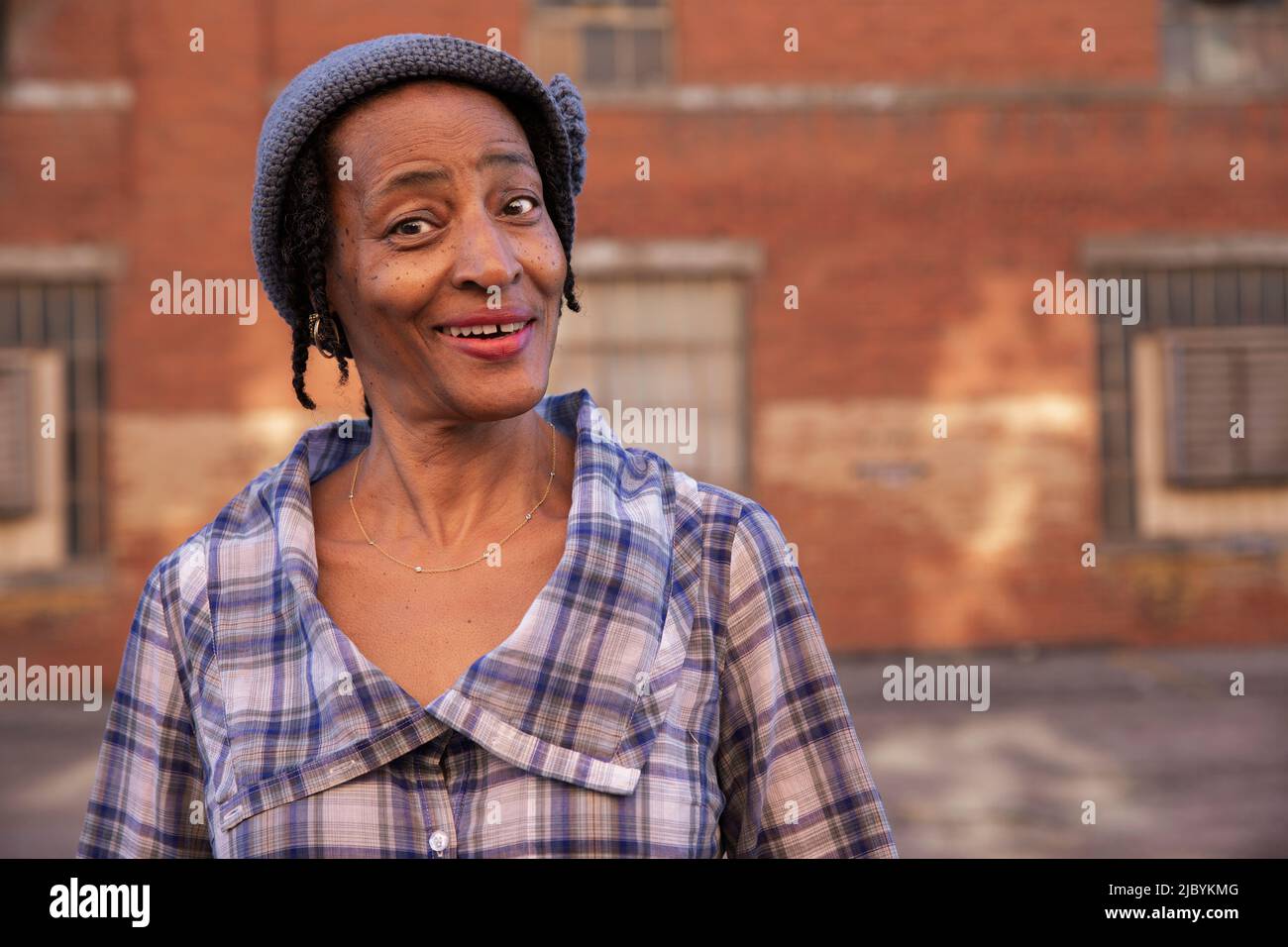 Portrait of older woman wearing knit hat standing in alley smiling looking at camera, brick wall in background Stock Photo