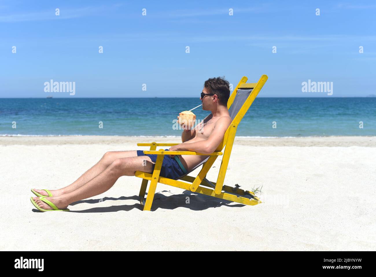 Young happy man drinks coconut juice sitting on deck chair on a south china sea beach in vietnam Stock Photo