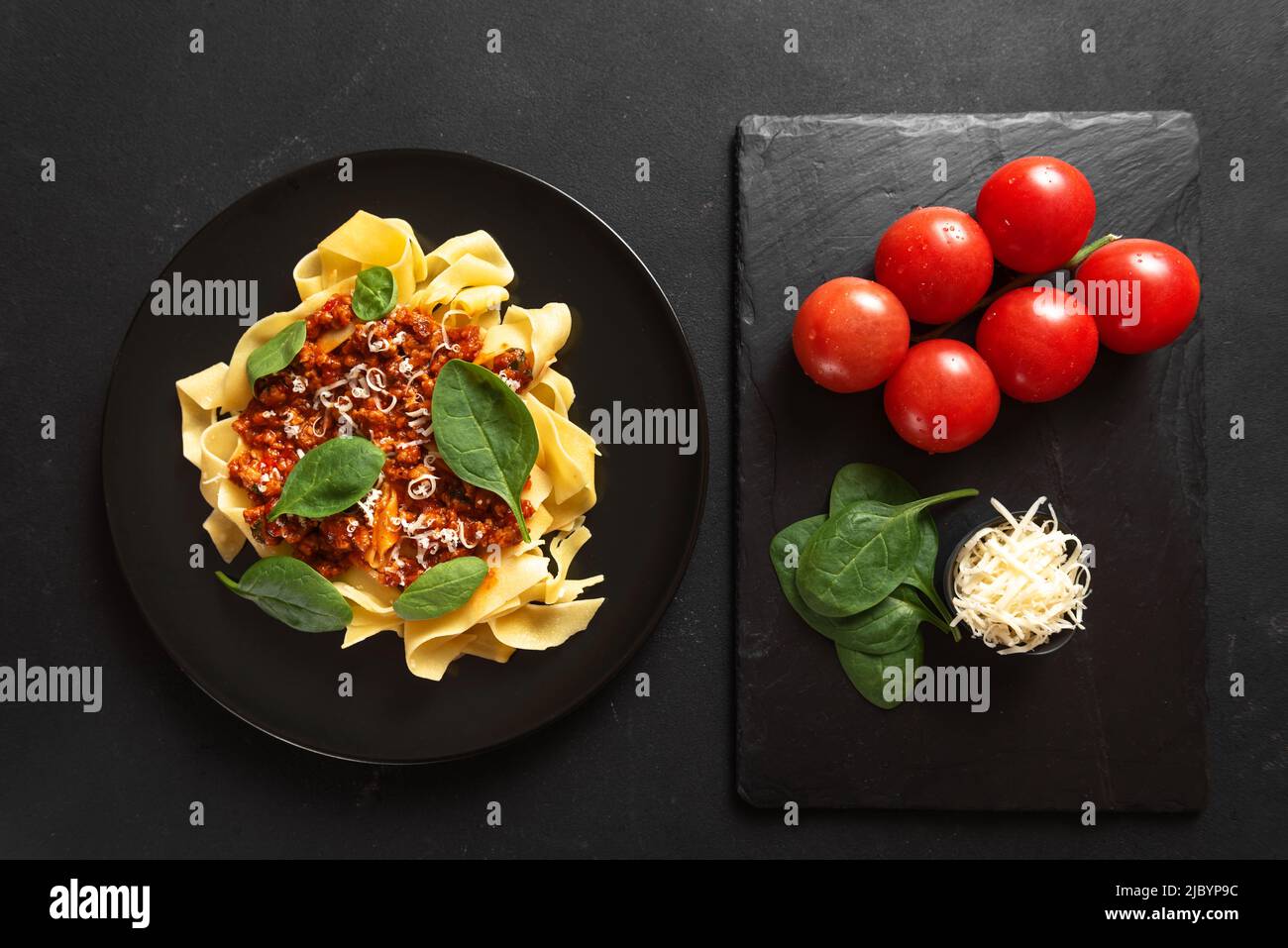 Traditional Italian pasta with cheese and basil on a black plate on a dark background Next to the ingredients for cooking tomatoes, basil and cheese Stock Photo