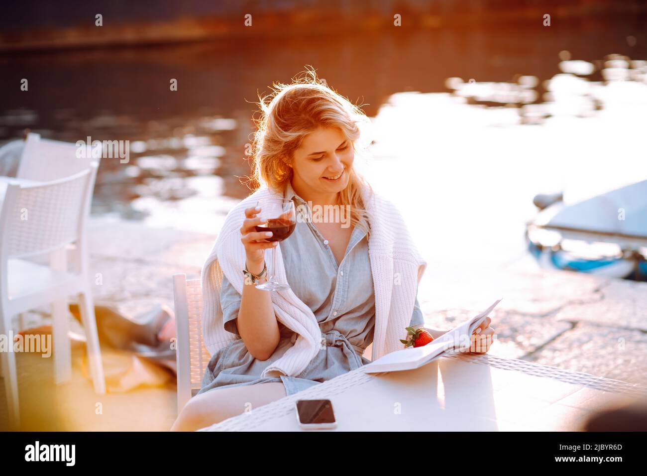 Young smiling attractive woman sitting at table, holding glass of red wine, reading book on embankment at sunset. Stock Photo