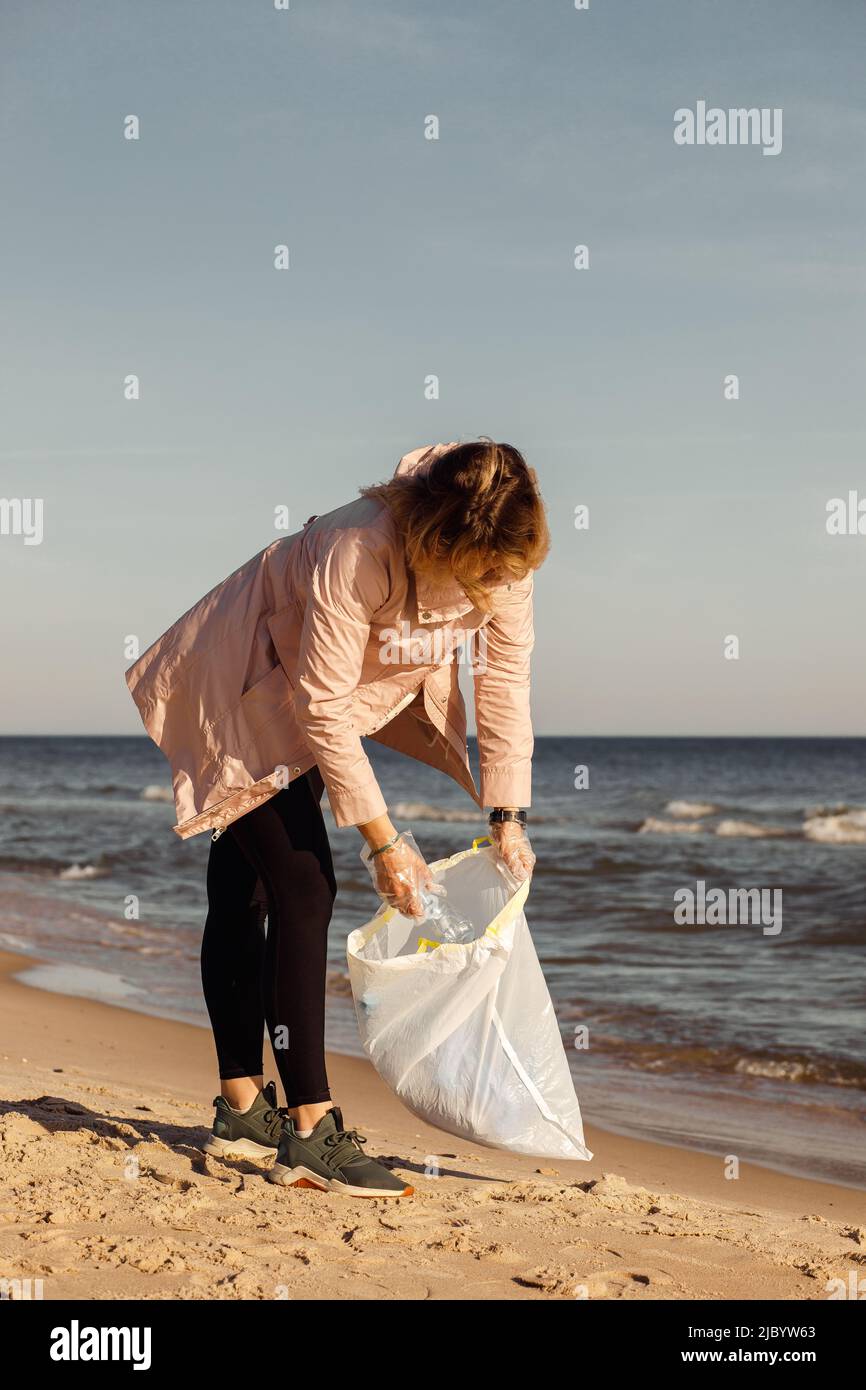 Vertical woman volunteer and assistant helping cleaning sandy beach near sea, cololect plastic waste, garbage. Ecology Stock Photo