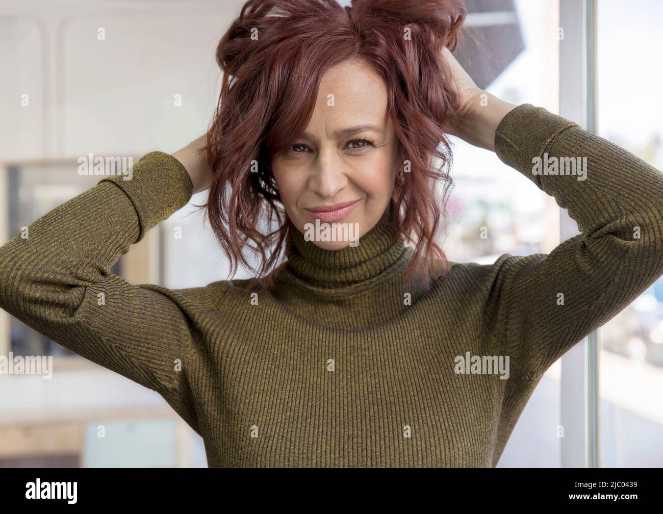 Portrait of a middle-aged woman looking into camera with both her hands up in her hair. Stock Photo