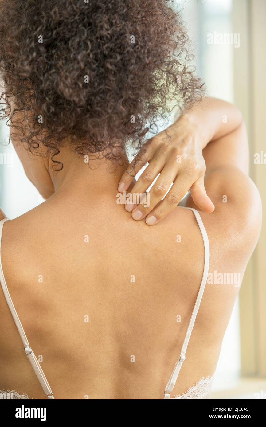 A middle-aged, mixed race woman with her back towards camera rubs her neck. Stock Photo