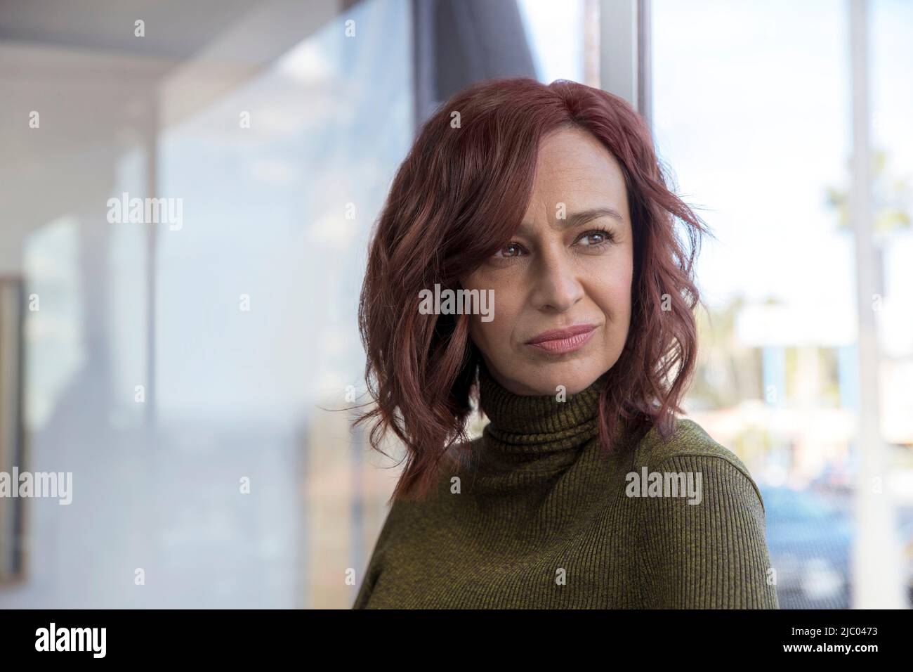 Mature woman with red hair, her back towards window, looks off frame. Stock Photo