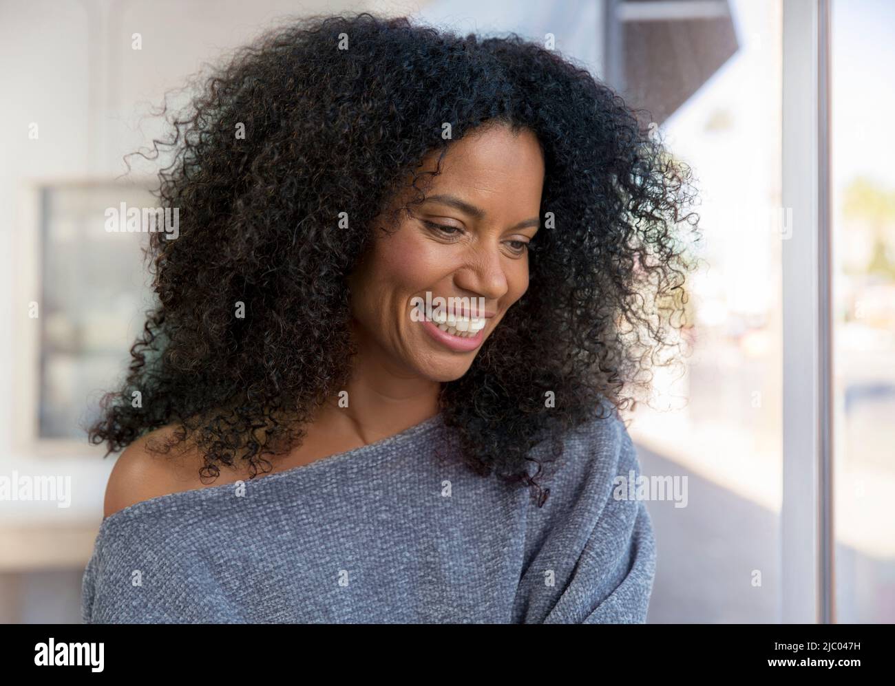 Mixed race, middle-aged woman with natural hair looking out window with a large smile. Stock Photo