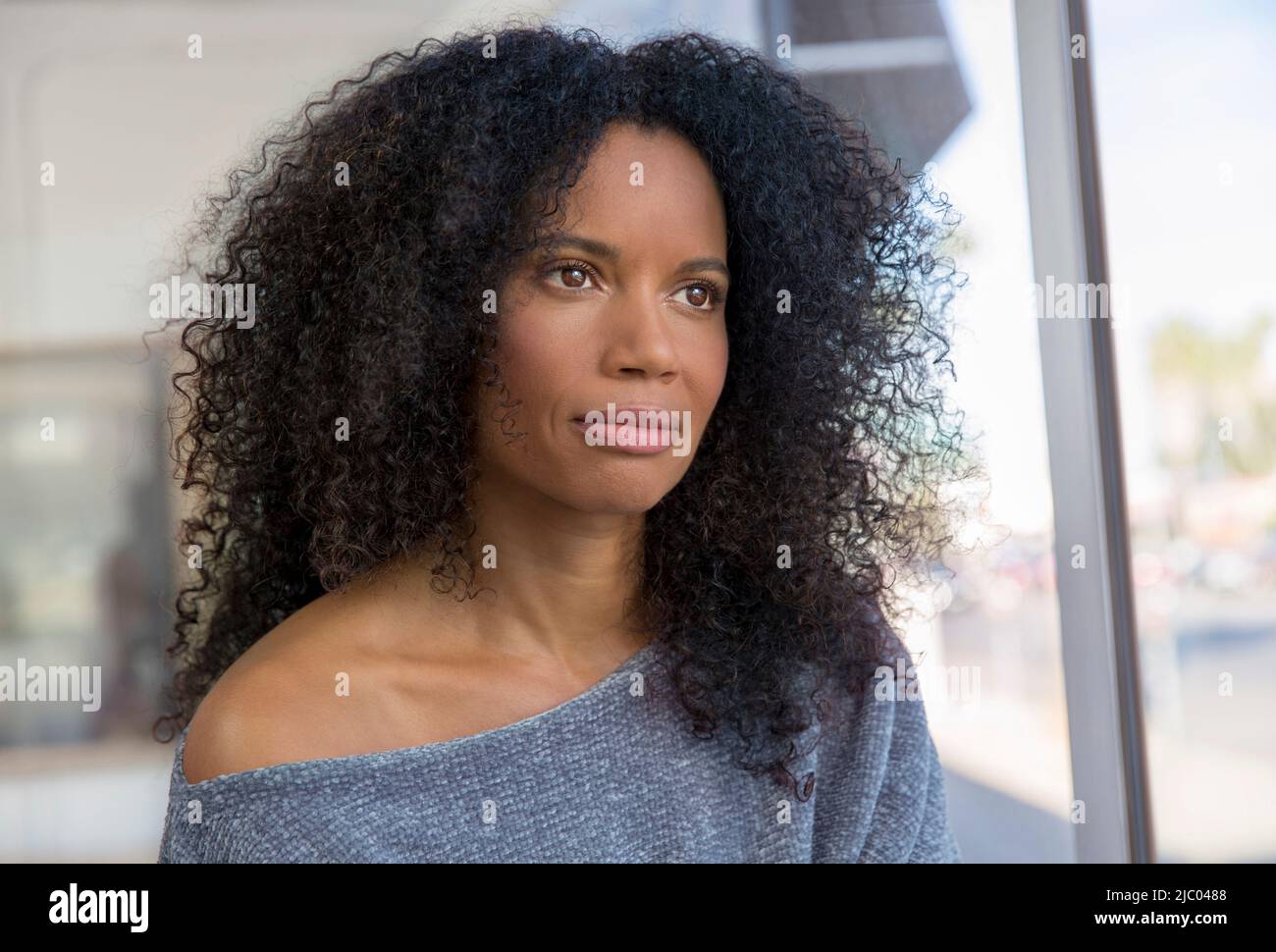 Mixed race, middle-aged woman with natural hair looking out window in brightly lit room. Stock Photo