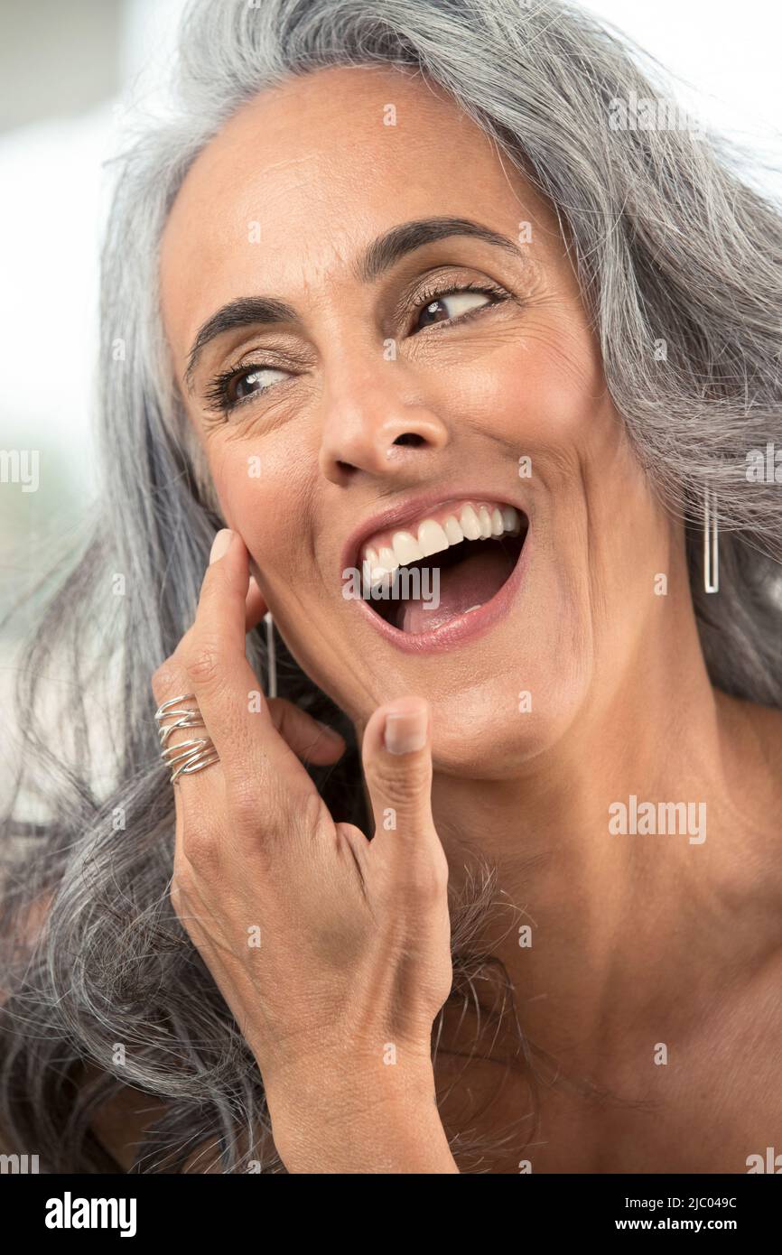 Middle-aged woman with gray hair brushes hair out of her face and looks off camera laughing. Stock Photo