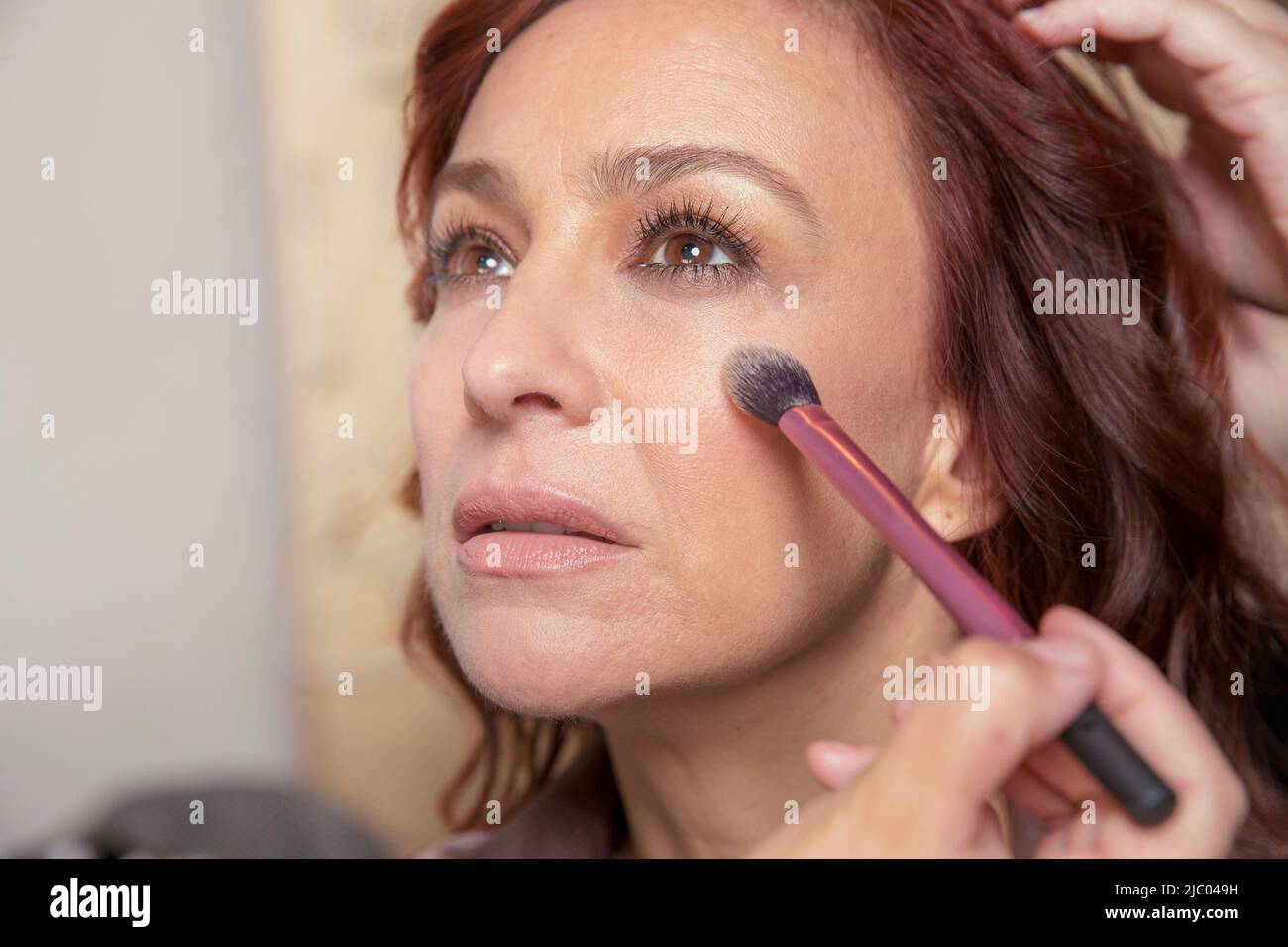 A middle-aged woman looks straight ahead while makeup is applied to her face. Stock Photo