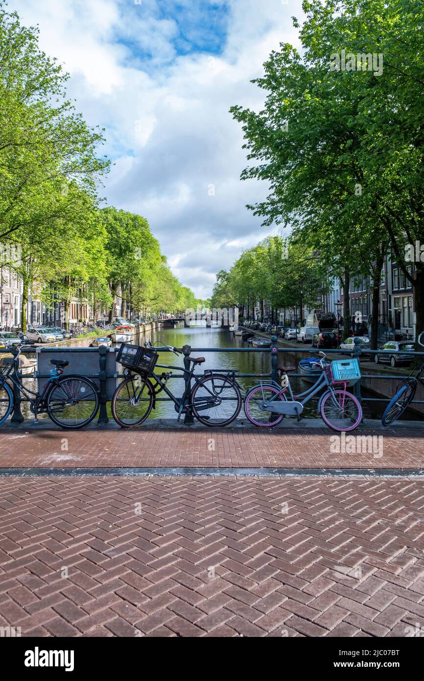 Bicycles parked on a bridge tiled floor over canal, moored boats on water. Amsterdam, Netherlands. Traditional houses brick facade and trees Stock Photo