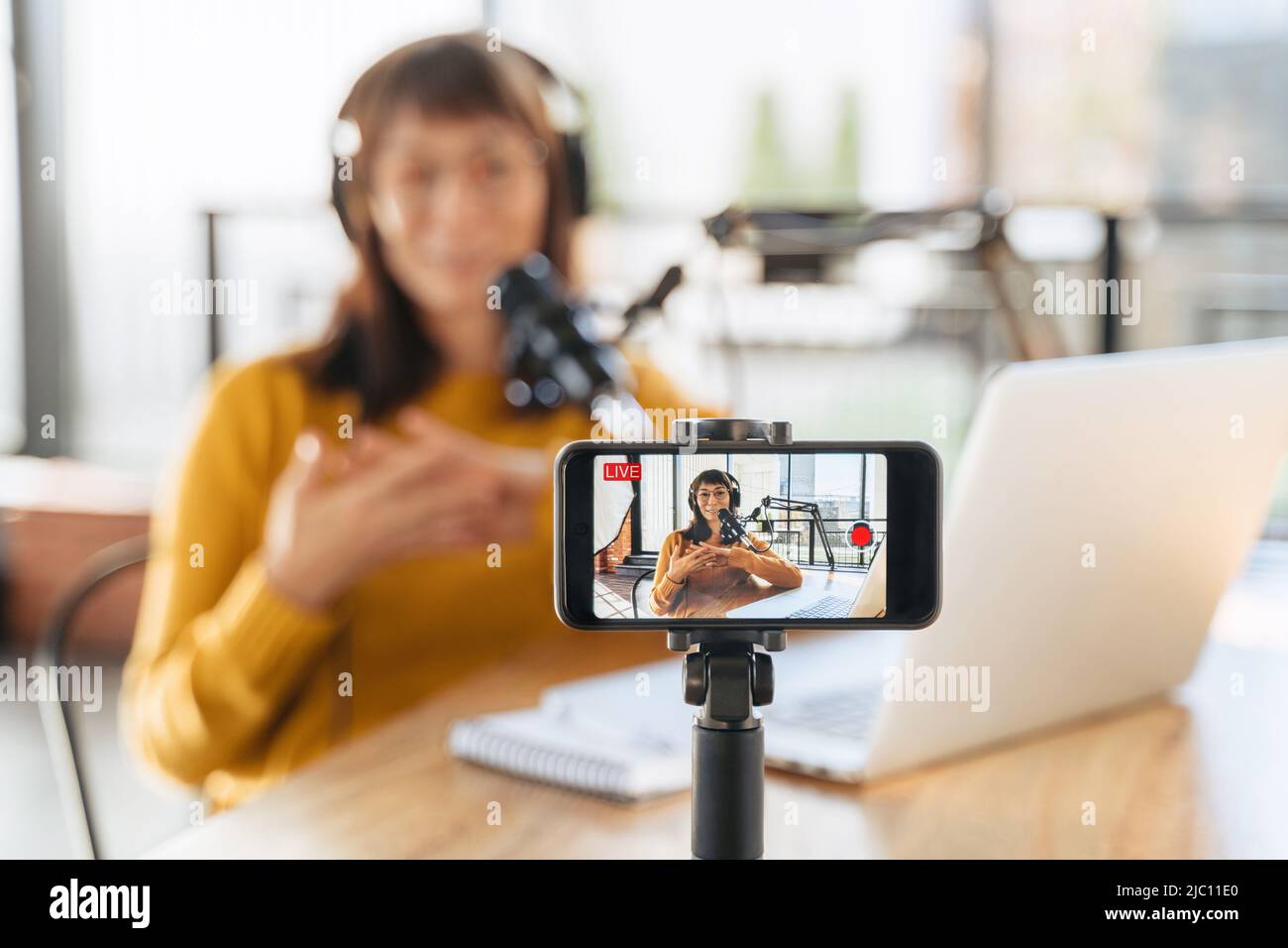Female vlogger live streaming podcast using microphone, laptop and cellphone on tripod. Selective focus on smartphone camera screen with woman podcaster recording and broadcasting live video. Close-up Stock Photo