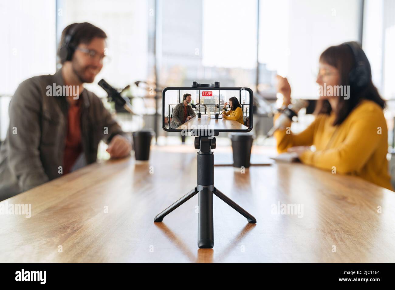 Man and woman hosts talking with each other while recording live video podcast in studio. Two podcasters in headphones shooting live video using smartphone on tripod. Selective focus on smartphone Stock Photo