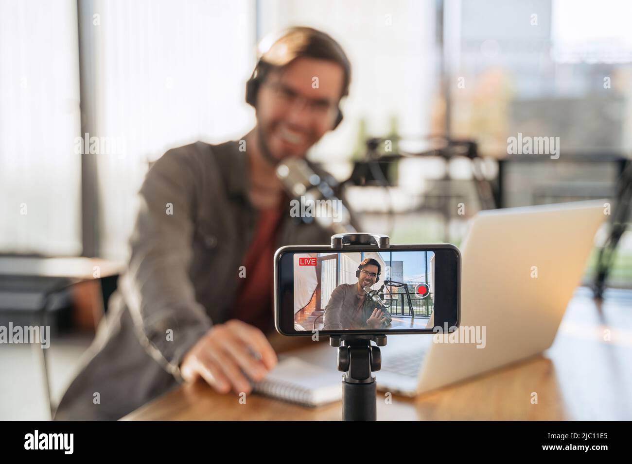 Male trendy podcast creator recording video podcast on smartphone. Selective focus on smartphone camera screen with man podcaster vlogger in headphones and glasses shooting live video for his channel Stock Photo