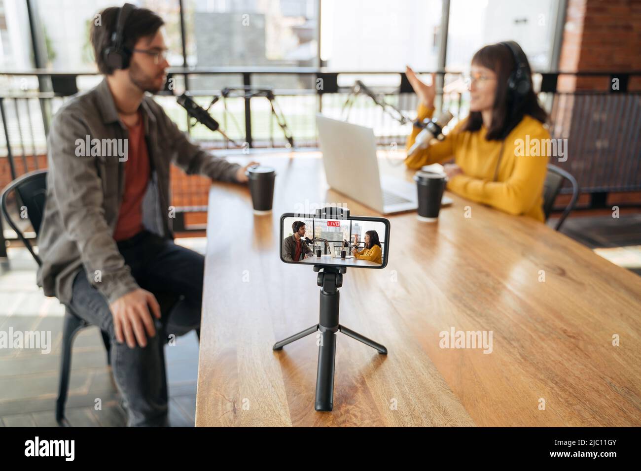 Two podcasters, man and woman talking while making live video podcast in studio, using microphones and headphones. Woman host streaming live video with man guest. Selective focus on smartphone screen Stock Photo